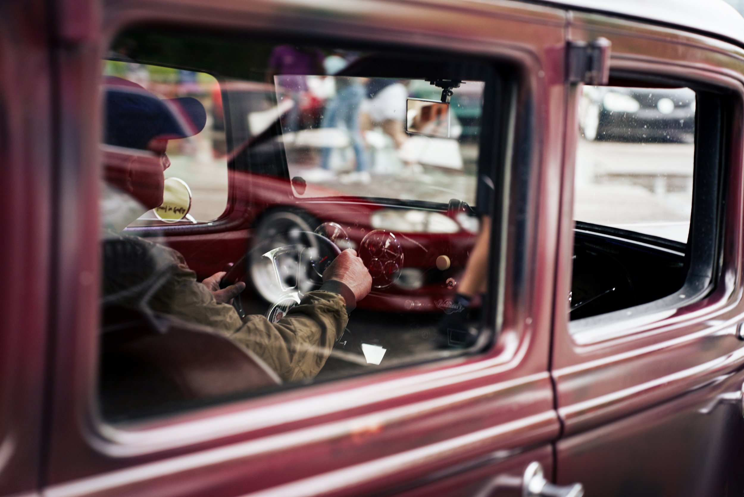 Close-up of a vintage red car's interior through the rear window, showing the driver wearing a cap and jacket, gripping the steering wheel with other blurry vehicles in the background.