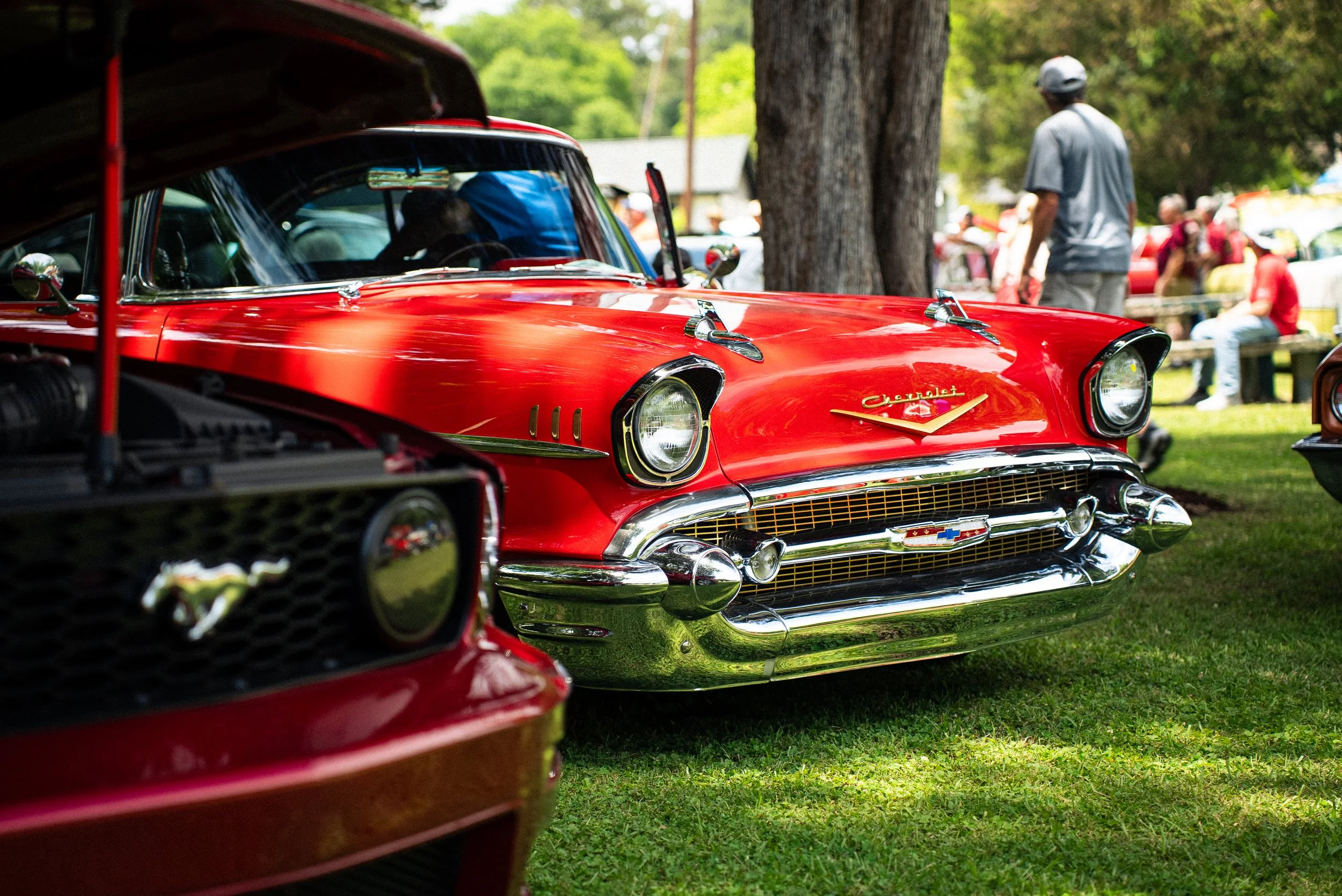 A close-up of a shiny red vintage Chevrolet car displayed outdoors at a car show, with presentation stands and people in the background.