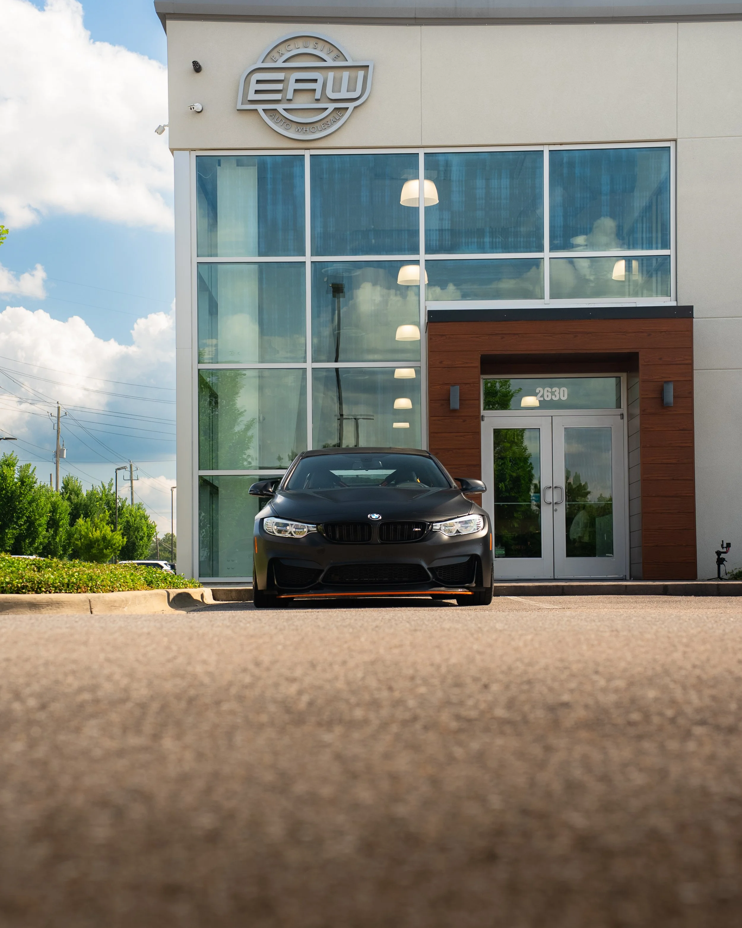 Front view of a black BMW car parked in front of a modern auto wholesale building with large glass windows and a wooden section displaying the number 2630.