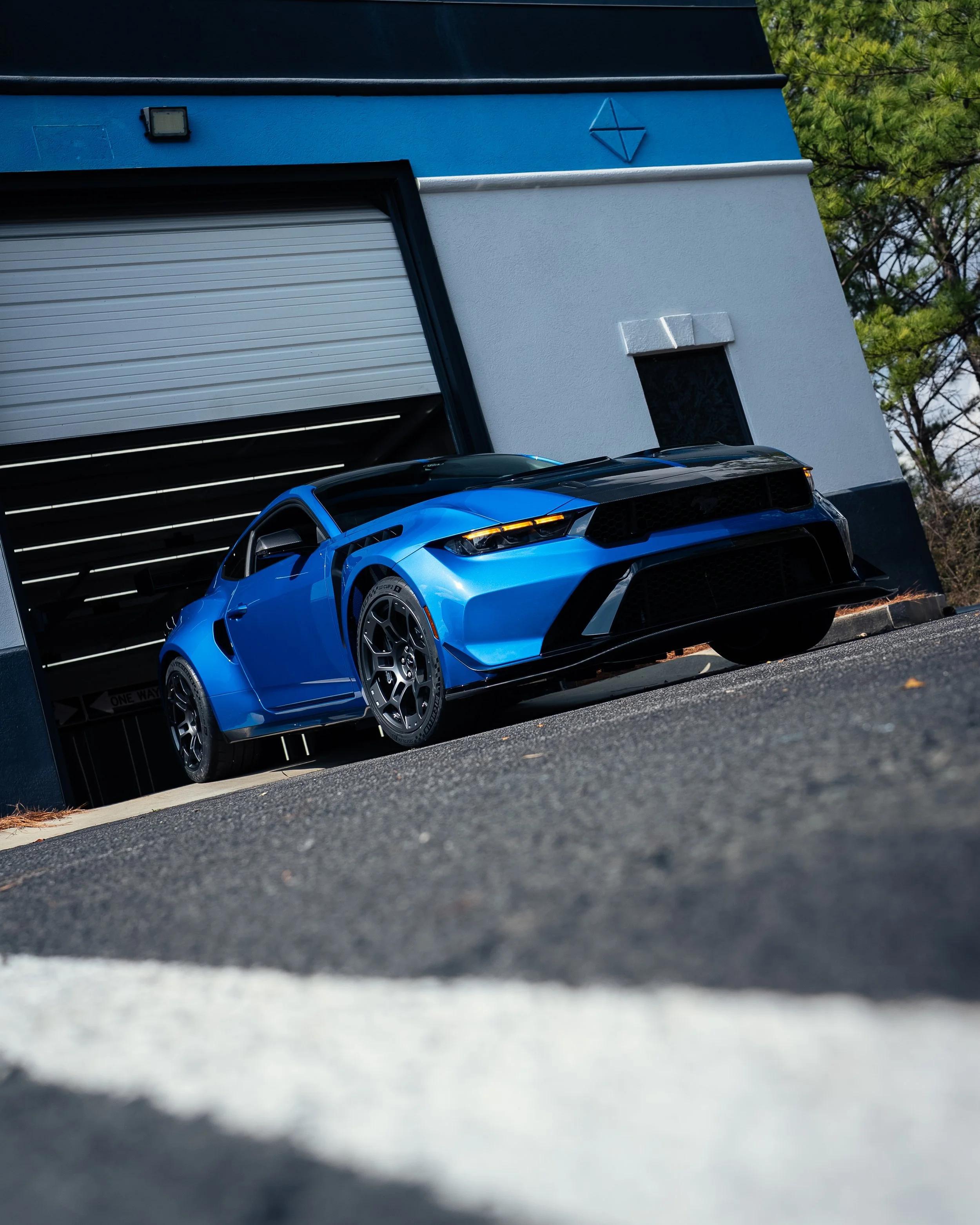 Blue sports car parked inside a building garage with a partly open gray rolling door, white and gray walls, and a small window.