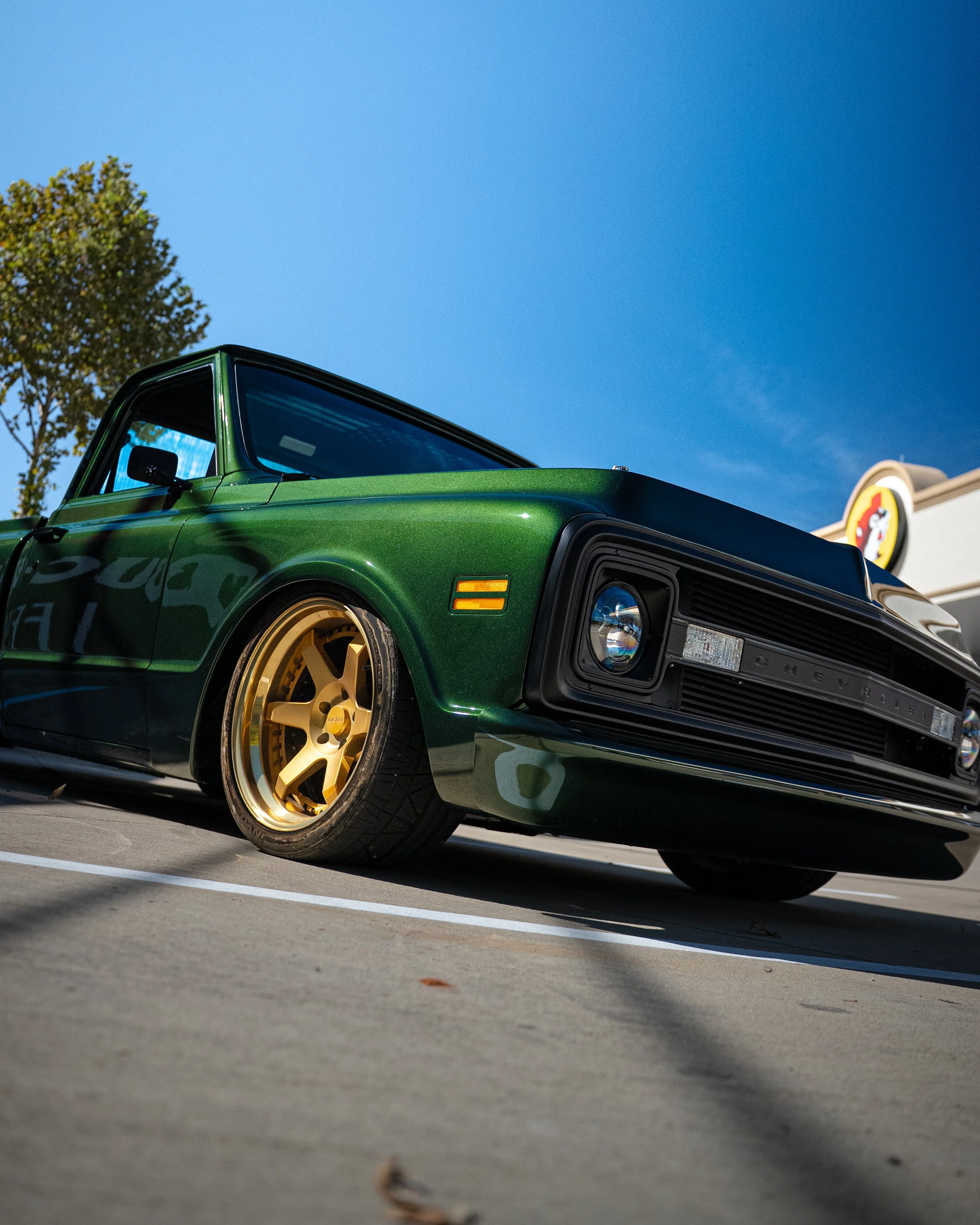 Close-up of a green classic Chevrolet pickup truck with gold wheels parked on a concrete surface, under a clear blue sky with a tree in the background and a Taco Bell sign on the right side.