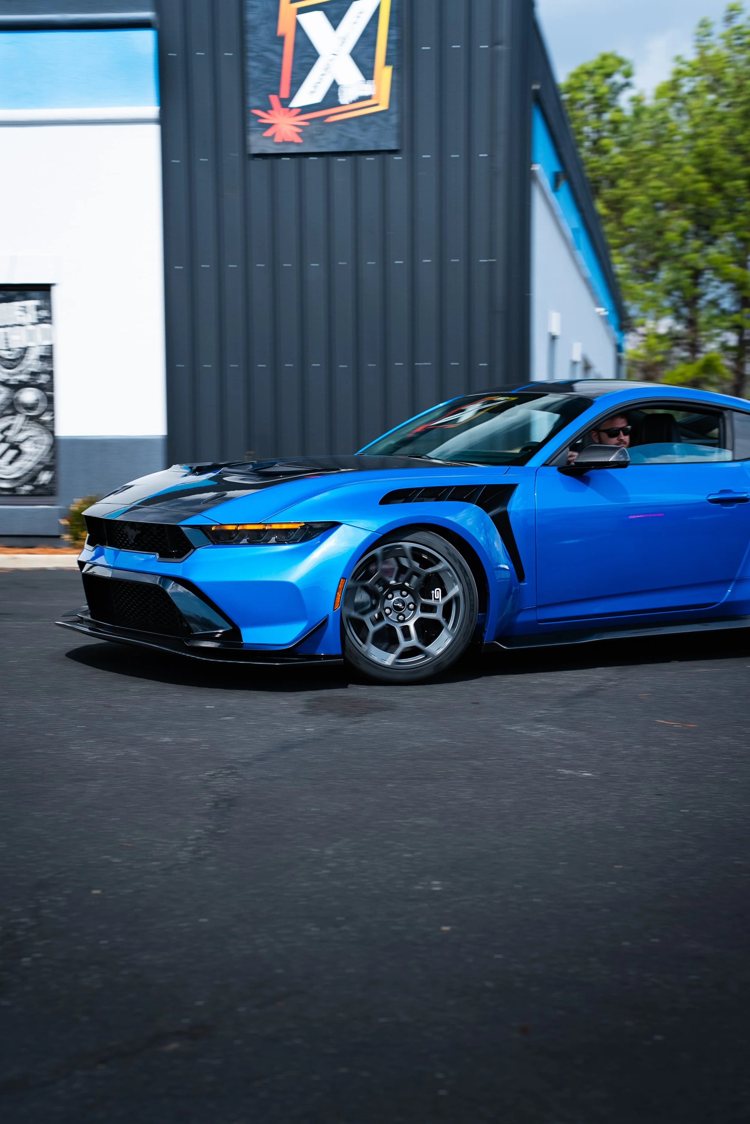 A blue Mustang GTD sports car with black accents driving on a street, with a man wearing sunglasses in the driver's seat and trees and a commercial building with an 'X' sign in the background.