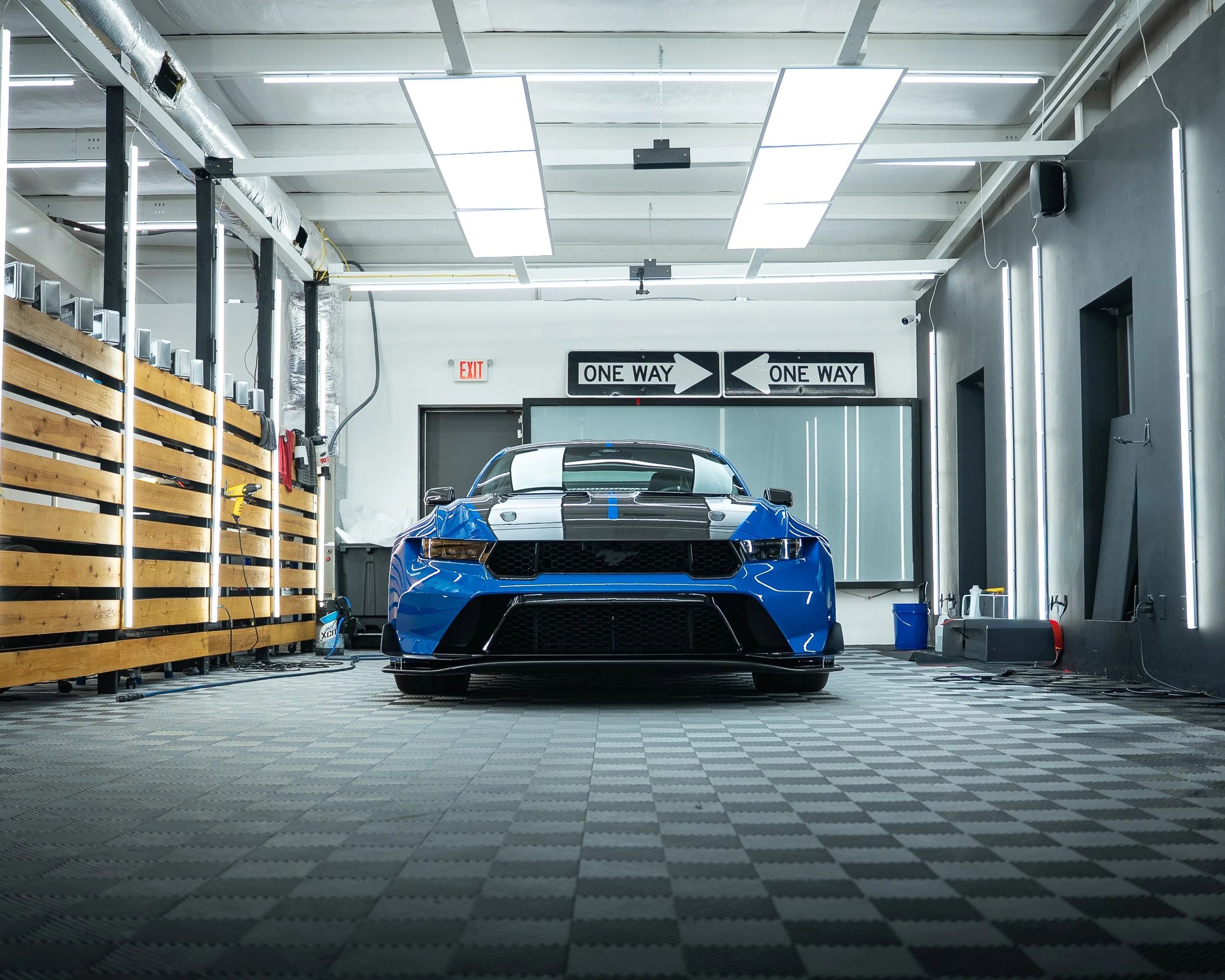 A blue sports car in a garage with a black and white checkered floor, shelves with tools, and bright overhead lighting.