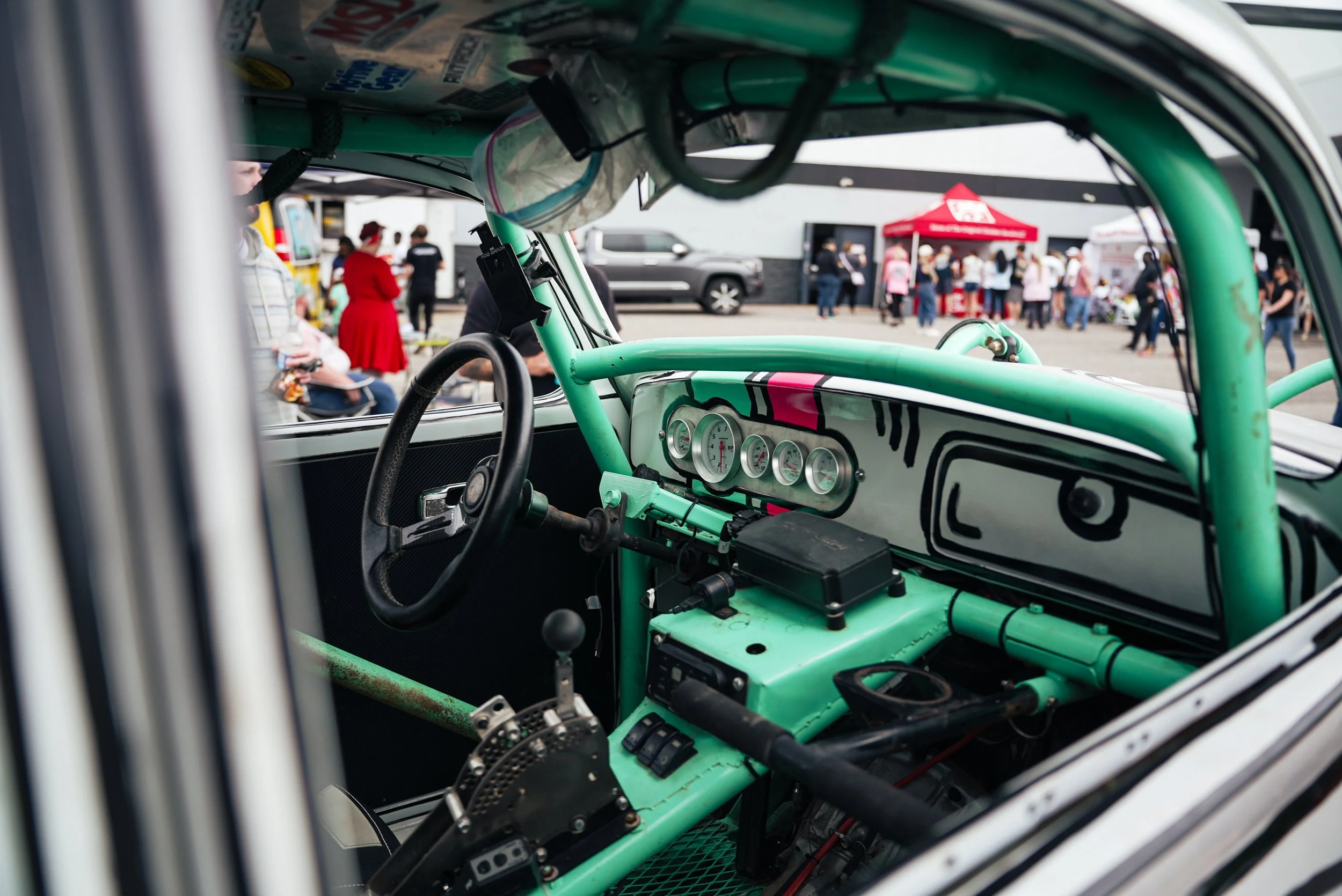 View from inside a race car focusing on the green dashboard with gauges, with a busy outdoor scene of people, tents, and vehicles seen through the windshield.