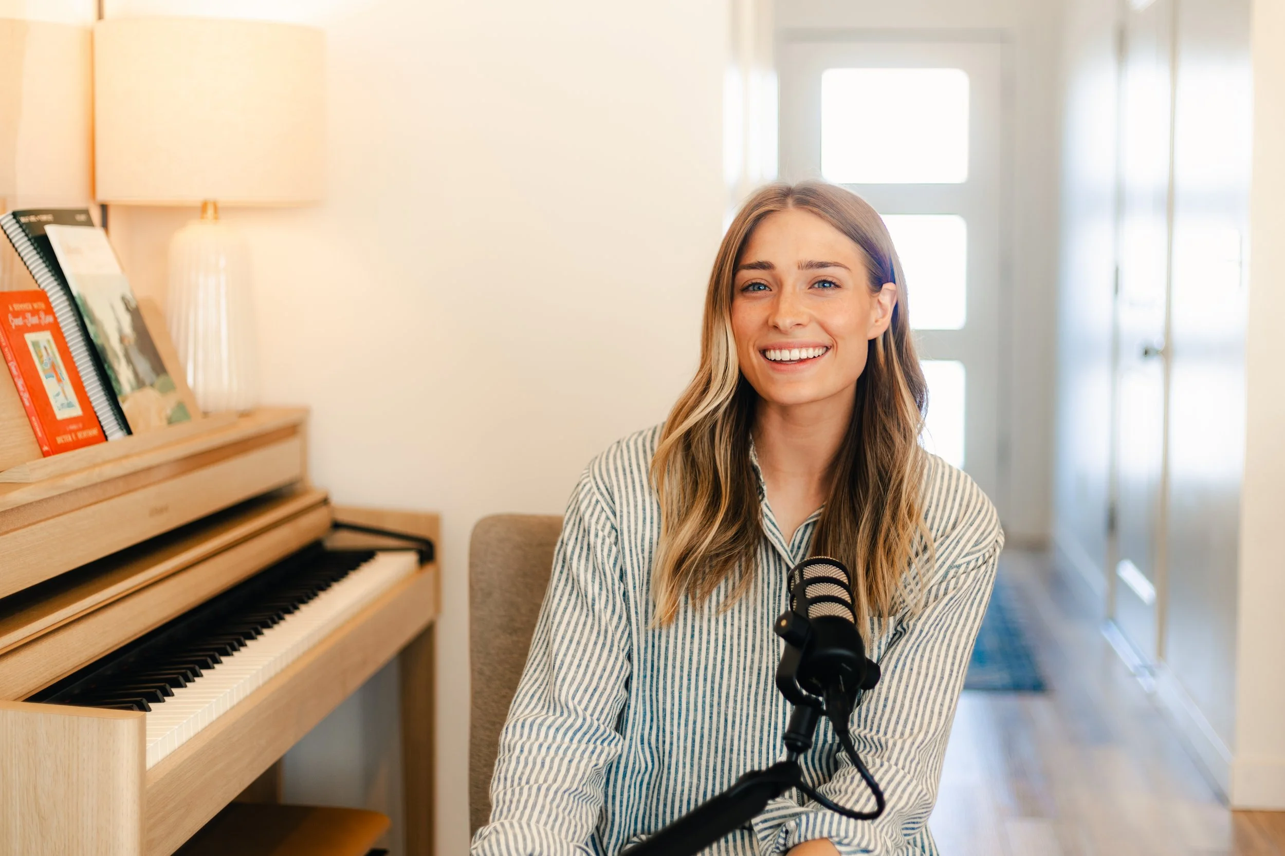 A young woman with long blonde hair smiling while sitting at a piano with a microphone in front of her, in a well-lit room.