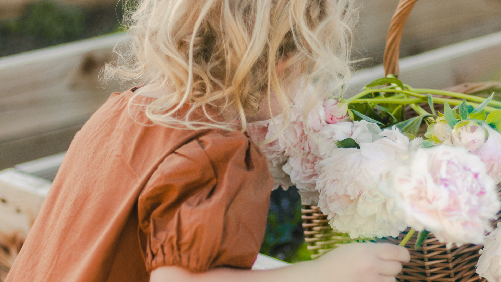 A young girl with curly blonde hair arranging pink and white peonies in a wicker basket.