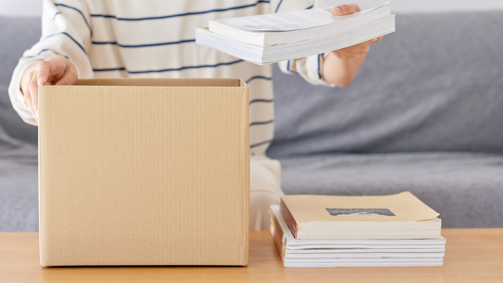Person packing books into a cardboard box with a gray sofa in the background.