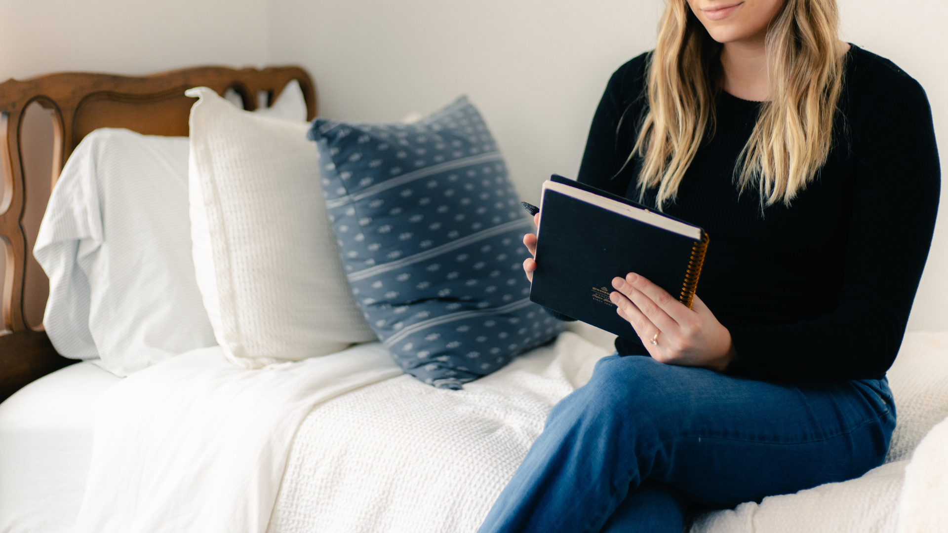 A woman sitting on a bed, holding a black notebook and pen, with pillows and a wooden headboard in the background.