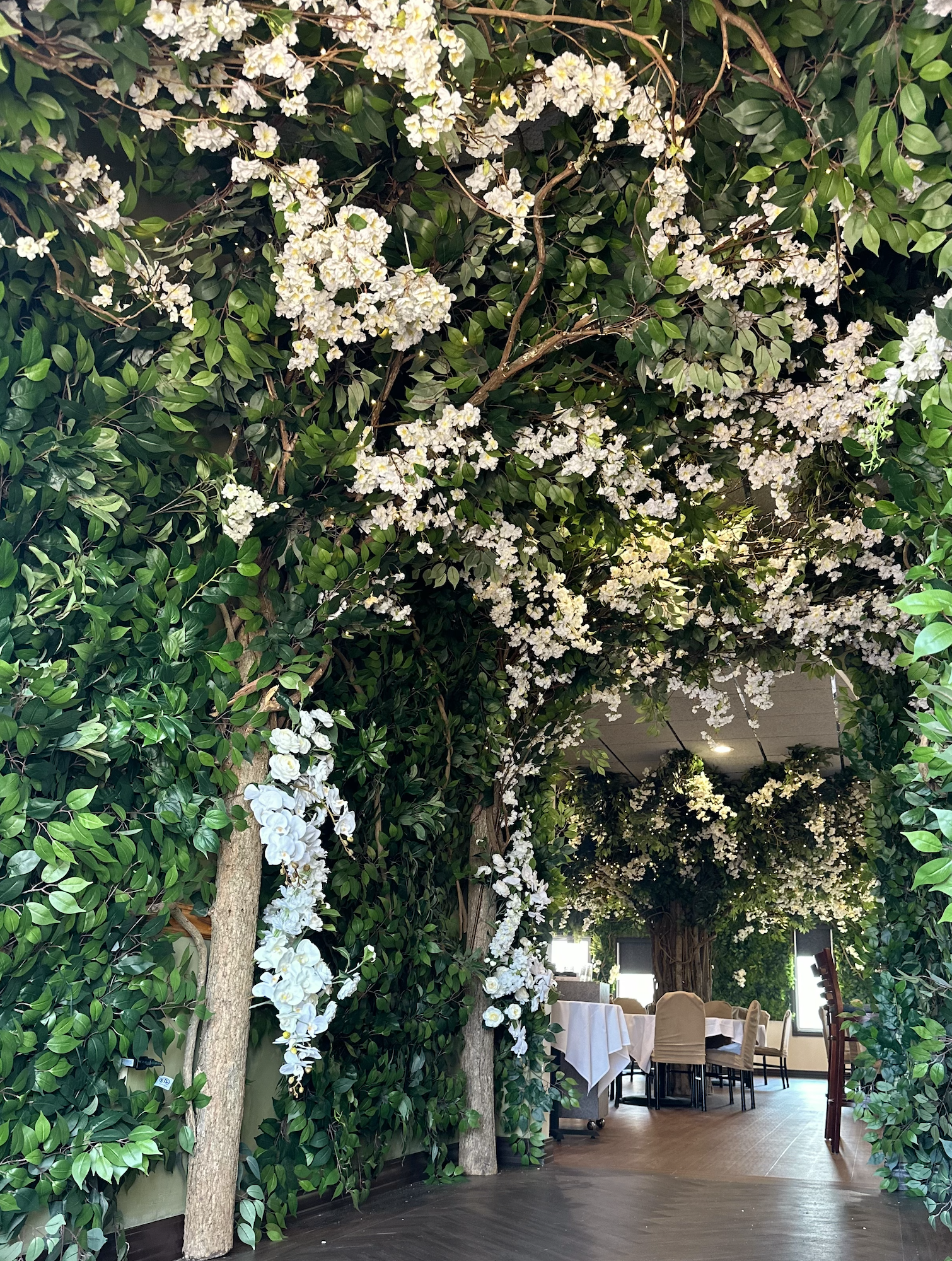 An indoor dining area decorated with an arch and ceiling covered in lush green leaves and white flowers.