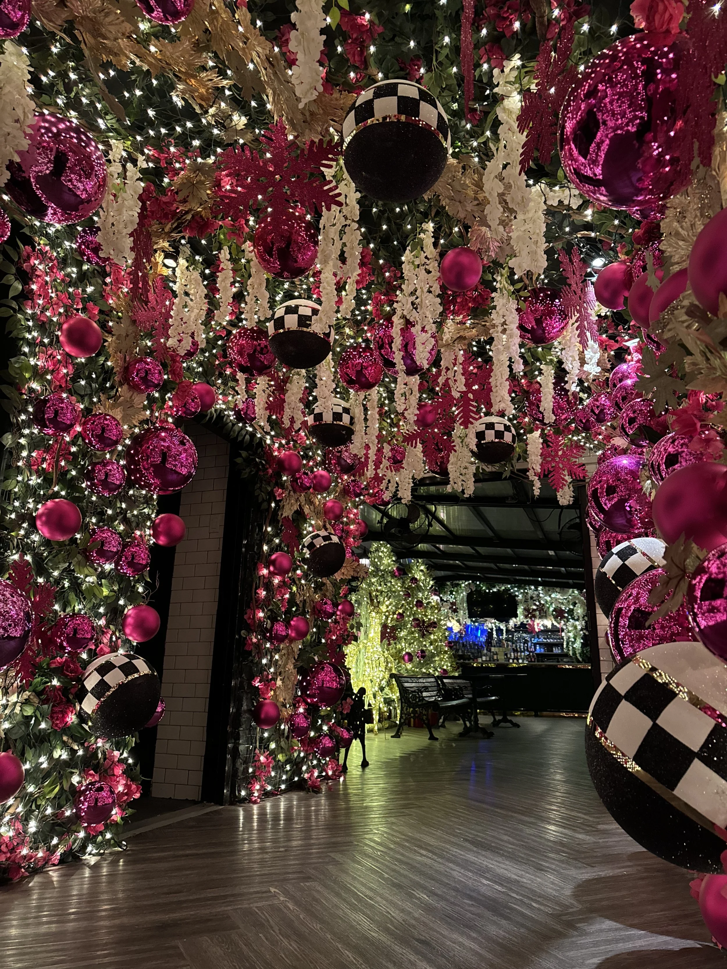 Festively decorated indoor space with pink, white, and black Christmas ornaments and hanging snowflakes, illuminated by string lights. A decorated arch leads to a serene seating area with a Christmas tree in the background.