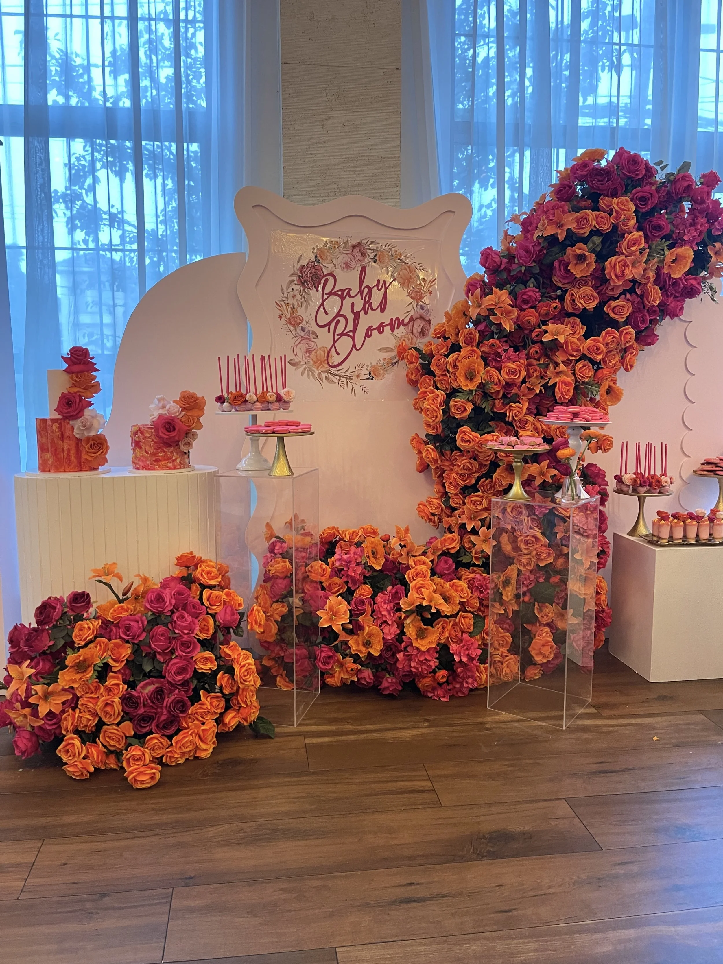 Decorative display with pink and orange roses, pink macarons, and pink candles, with a white backdrop featuring the words 'Baby Bloom' in pink, set against a wooden floor and blue-tinted windows.