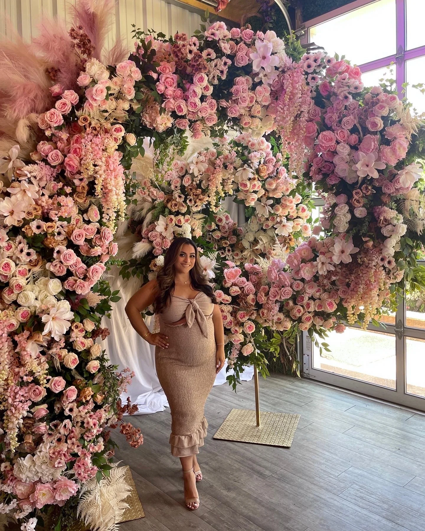 A woman in a beige dress standing in front of a large floral arrangement with pink, white, and purple flowers, inside a room with large windows.