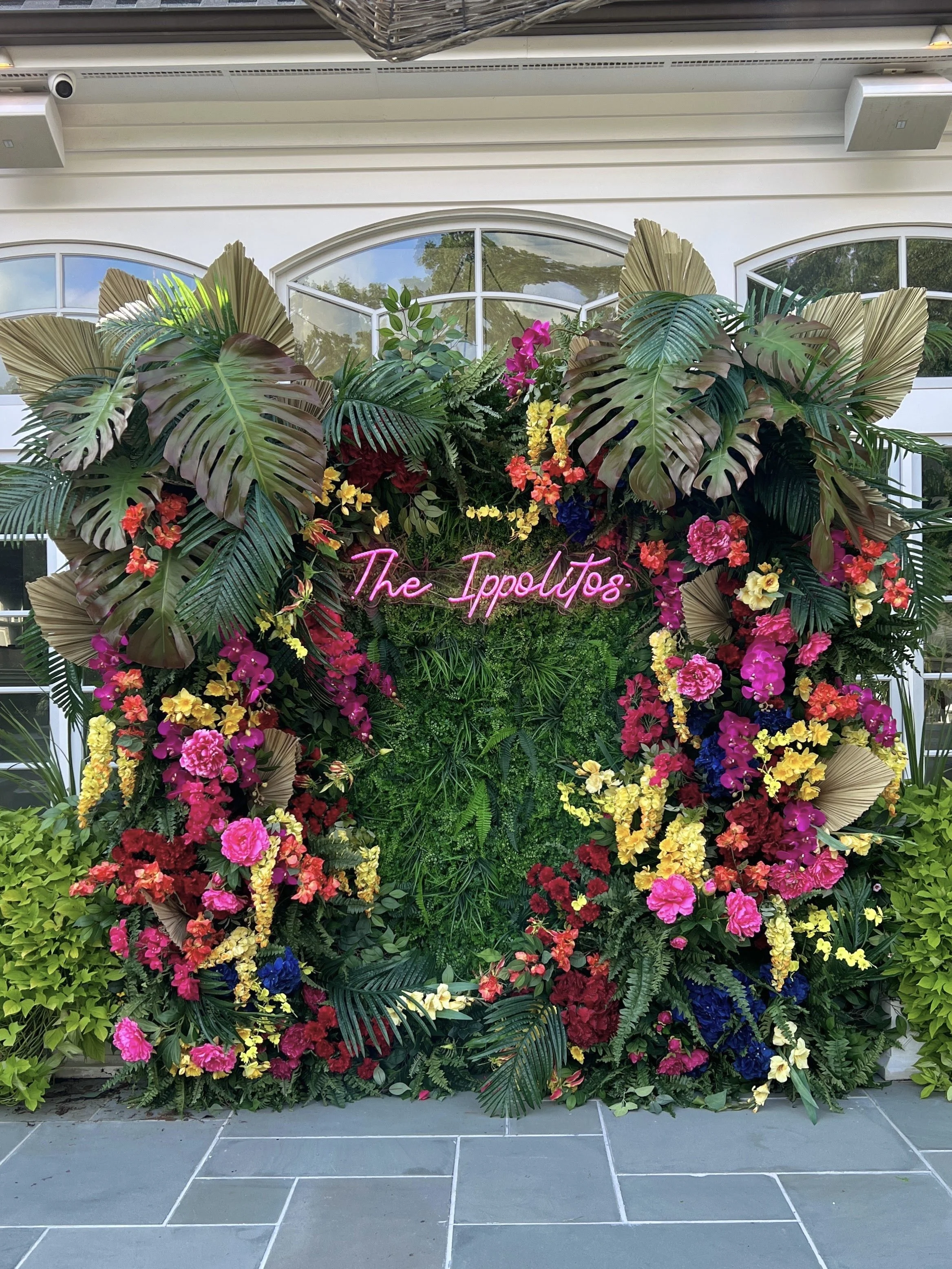 Flower arrangement with tropical leaves, colorful flowers, and a pink neon sign that reads 'The Ipoliots'