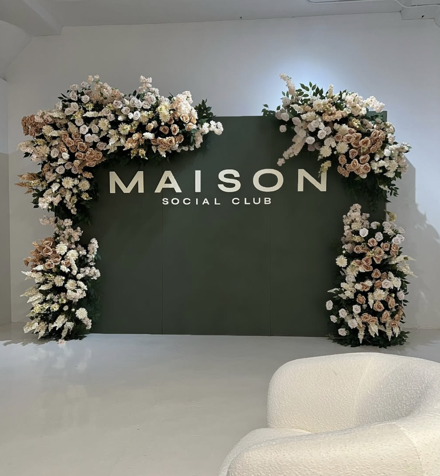 Green wall with white text reading 'MAISON SOCIAL CLUB' surrounded by elaborate arrangements of pink and white flowers.