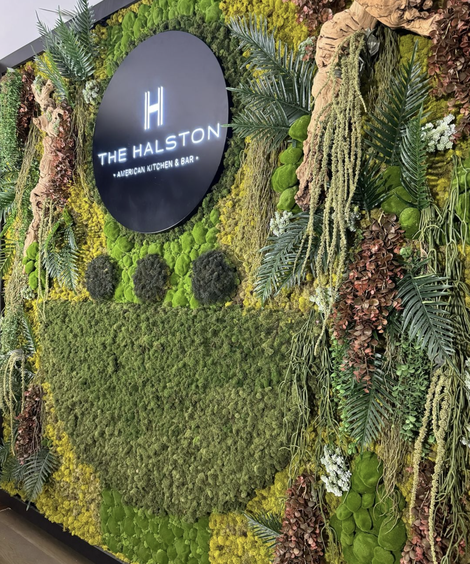 Decorative wall with green moss, plants, and flowers surrounding a round black sign with white neon text that reads 'The Halston' and 'American Kitchen & Bar'.