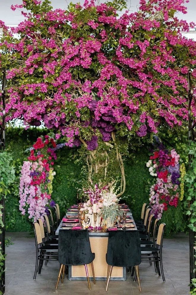 Elegant outdoor dining setup under a flowering pink bougainvillea tree with decorated tables and floral arrangements.