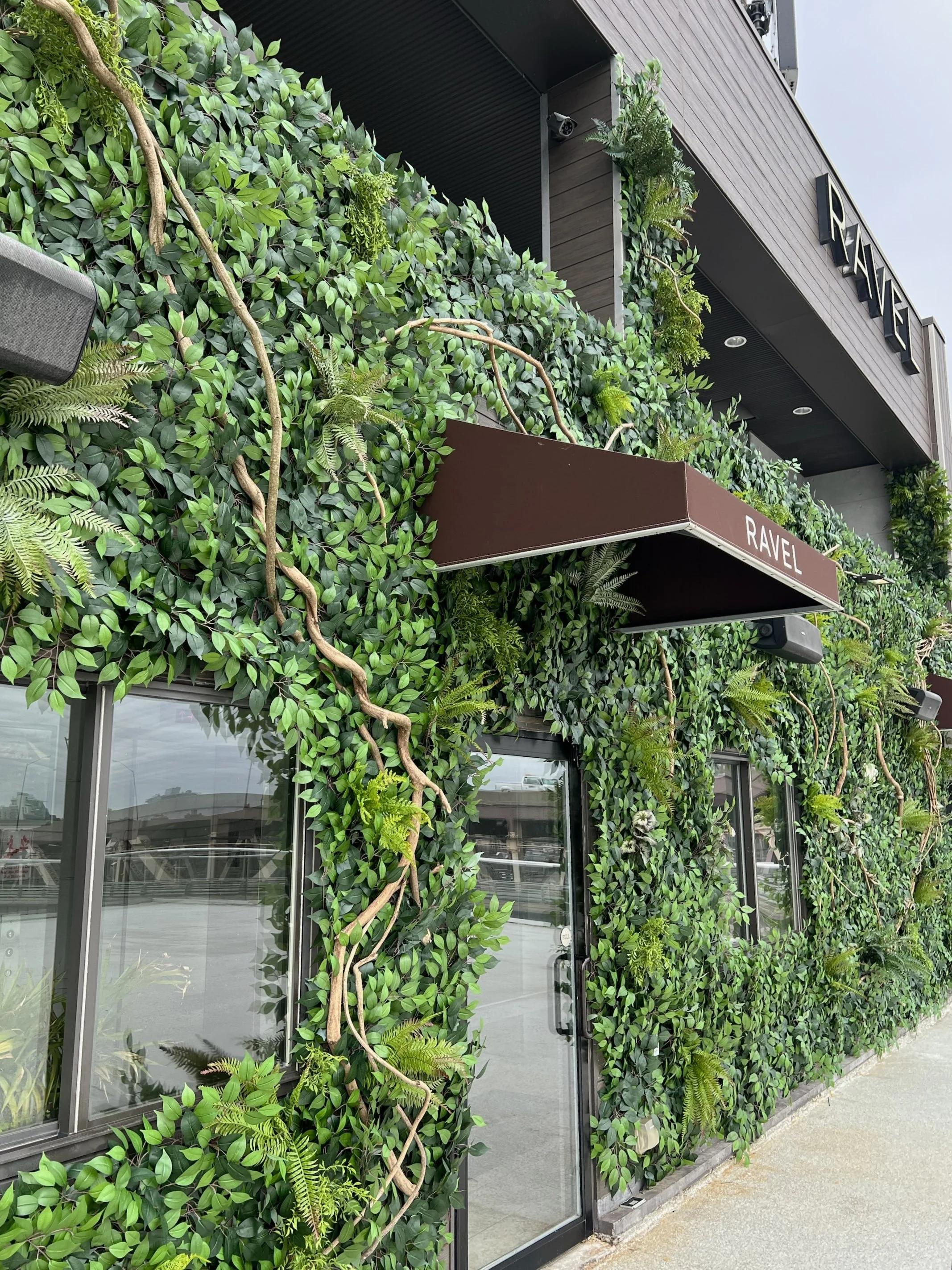 Exterior of a building covered in lush green foliage with windows and brown awnings labeled 'RAVEL'.