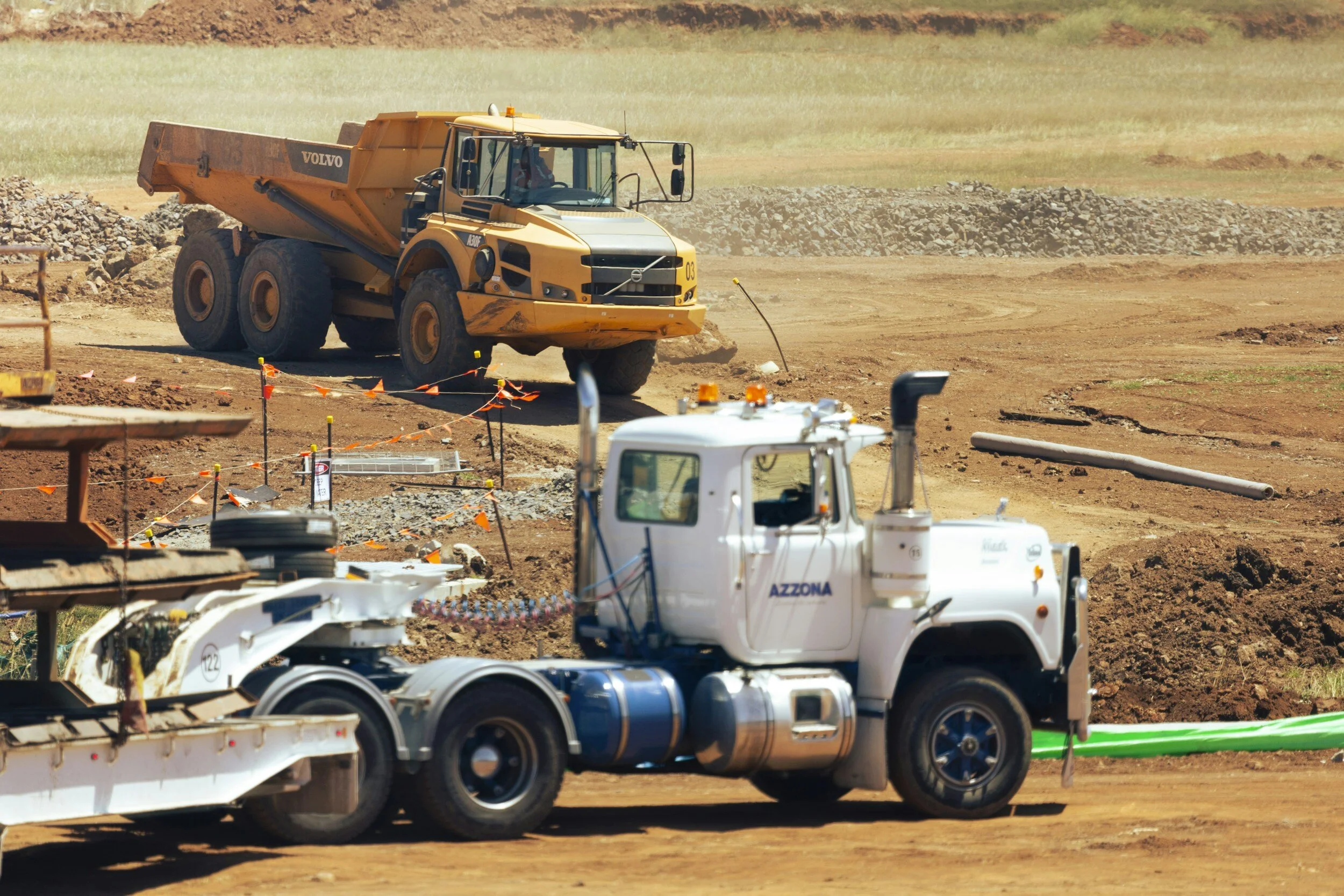 Construction site with a white truck labeled 'AZZONA' in the foreground and a yellow Volvo dump truck in the background on a dirt road.