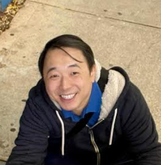 A man smiling and looking up, wearing a blue shirt and a black hoodie with a gray hoodie lining, outdoors on a sidewalk.