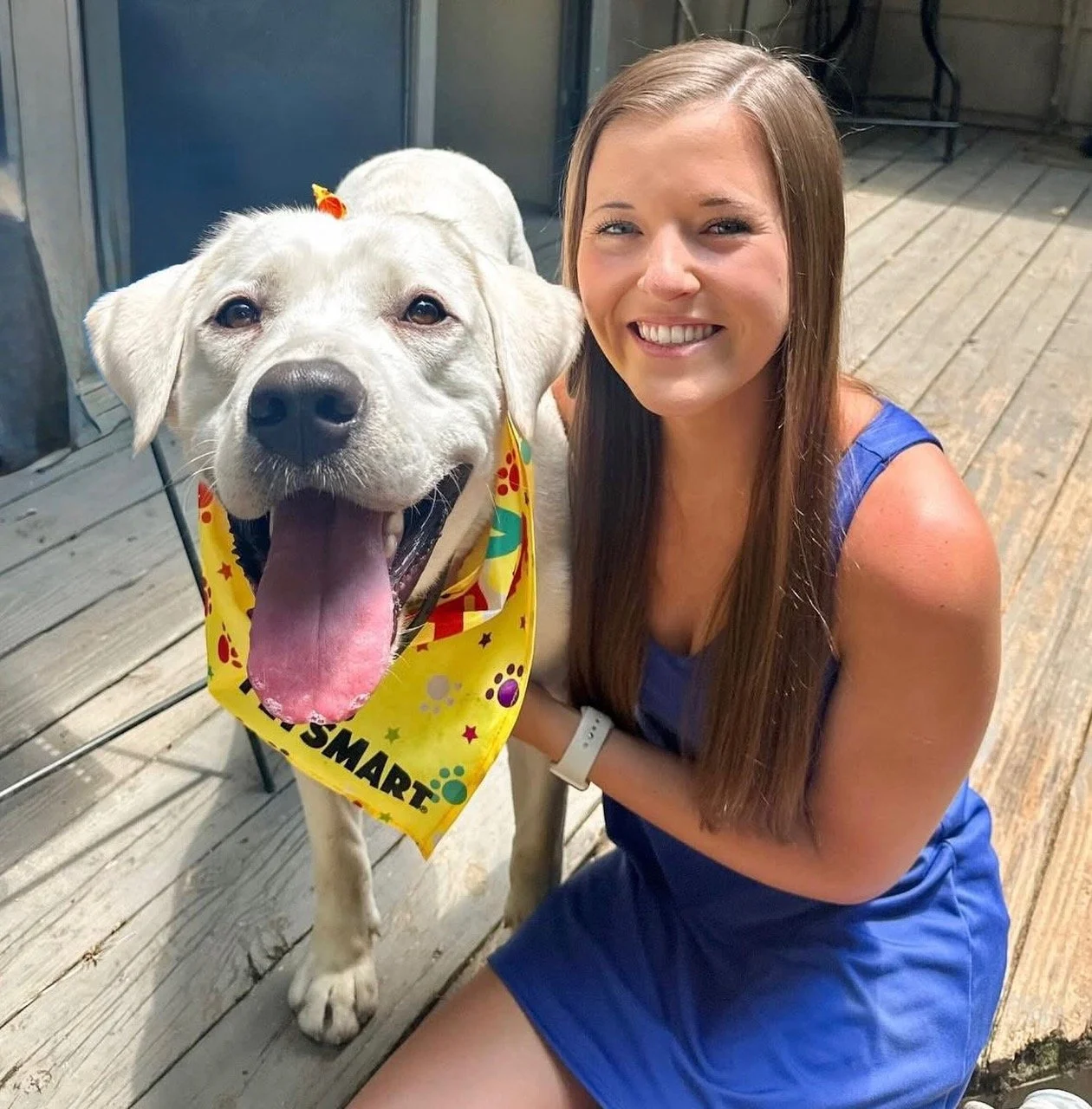 A woman sitting on a wooden deck, smiling next to a large yellow Labrador Retriever dog with a colorful bandana, both appearing happy.