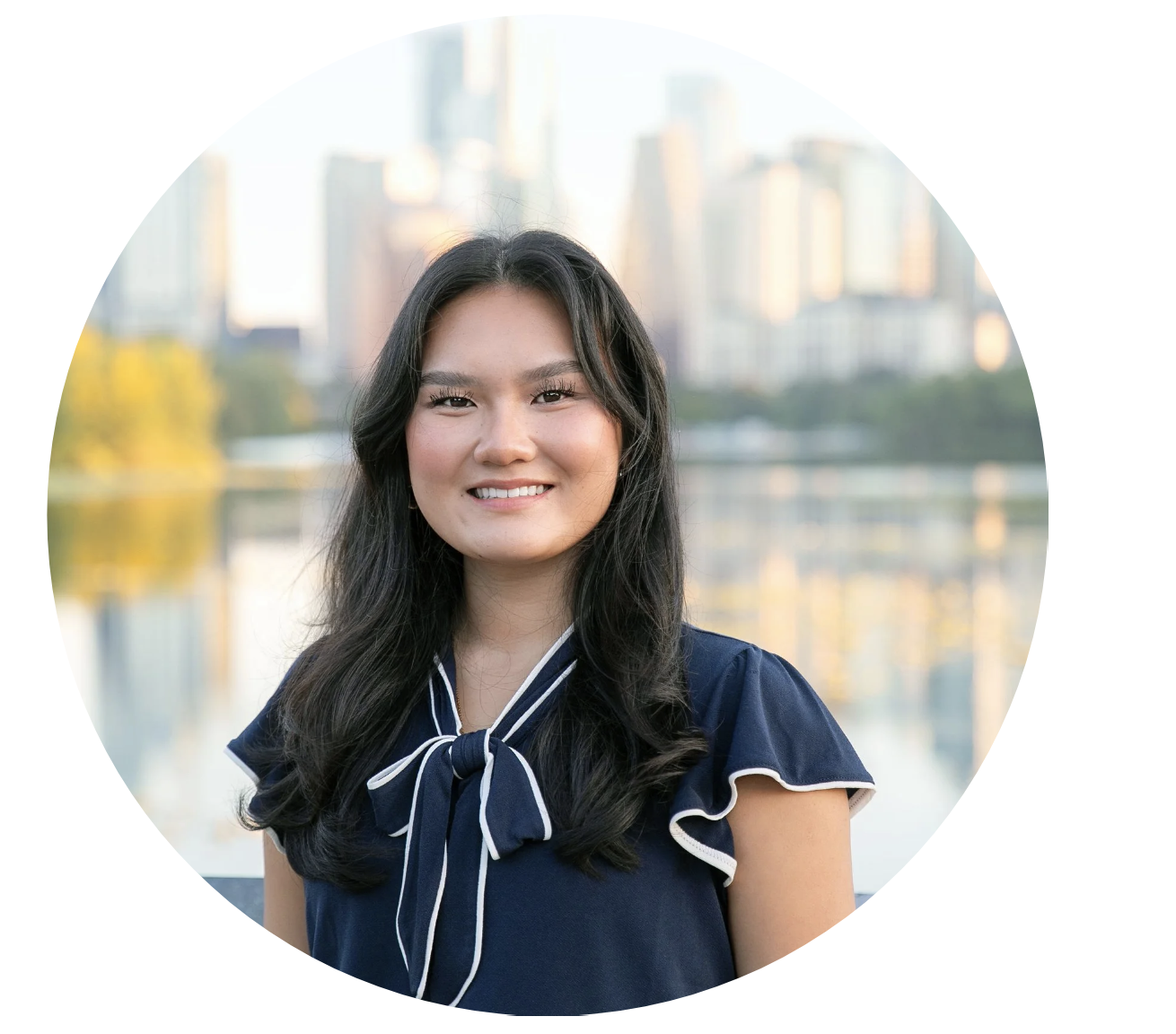 A young woman with long black hair smiling outdoors near a body of water with city skyline in the background