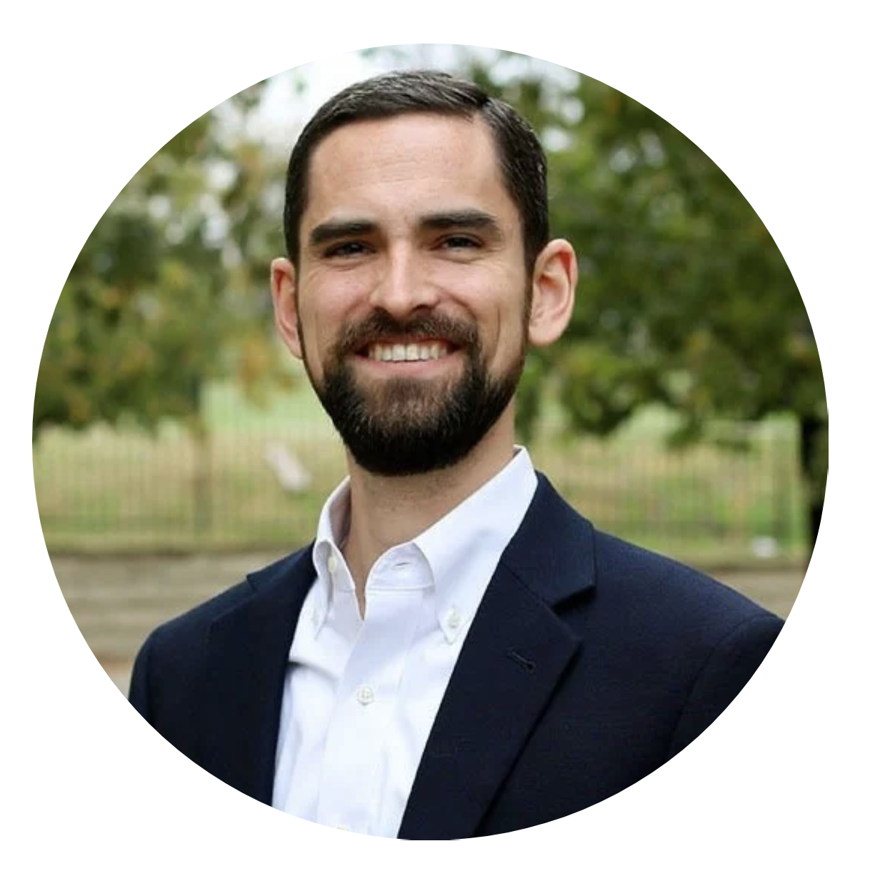 A man with dark hair, beard, and mustache, smiling, wearing a white shirt and a dark suit jacket, outdoors with trees in the background.