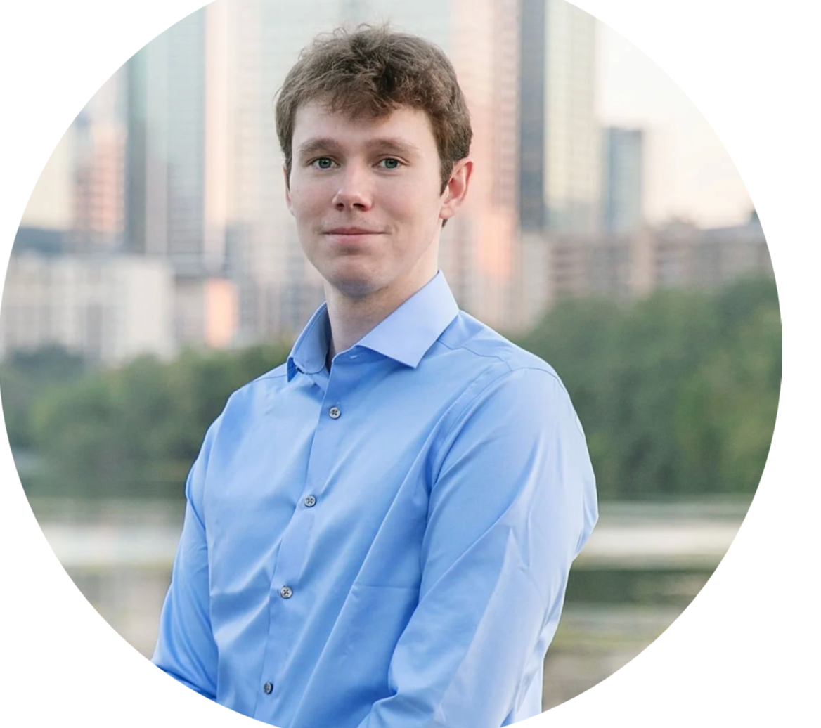A young man with light brown hair wearing a blue dress shirt, standing outdoors with a city skyline and trees in the background.
