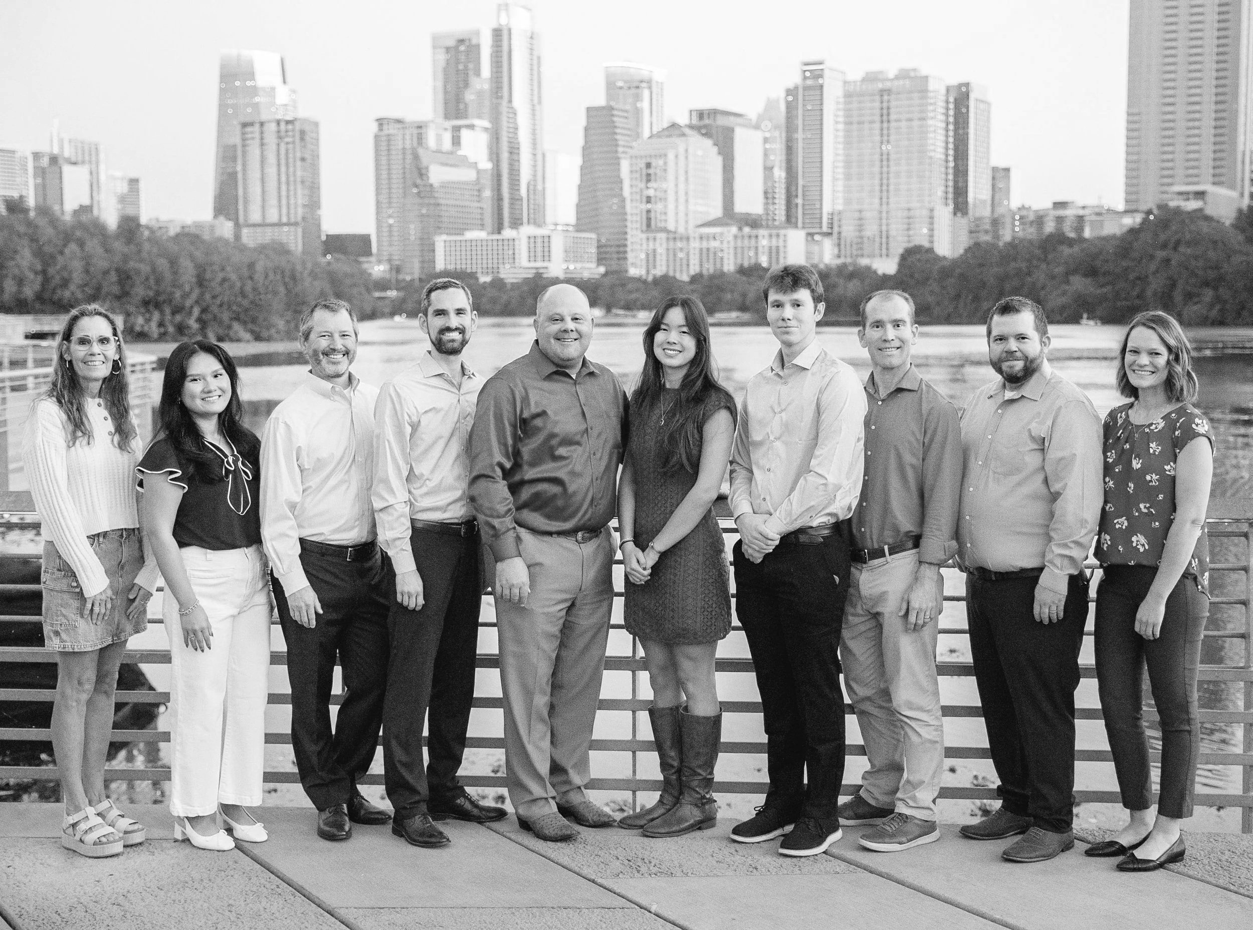 A diverse group of eleven people standing outdoors on a promenade with a city skyline in the background, smiling at the camera.