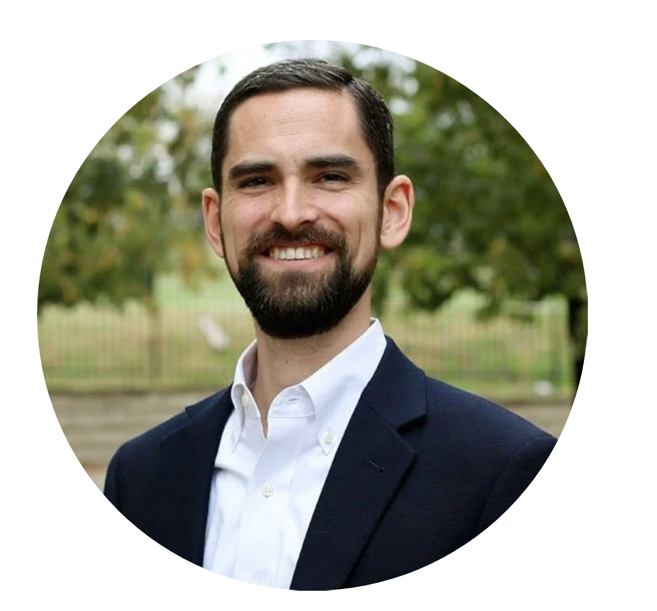 A smiling man with dark hair and a beard, wearing a white collared shirt and a dark blazer, standing outdoors with green trees in the background.