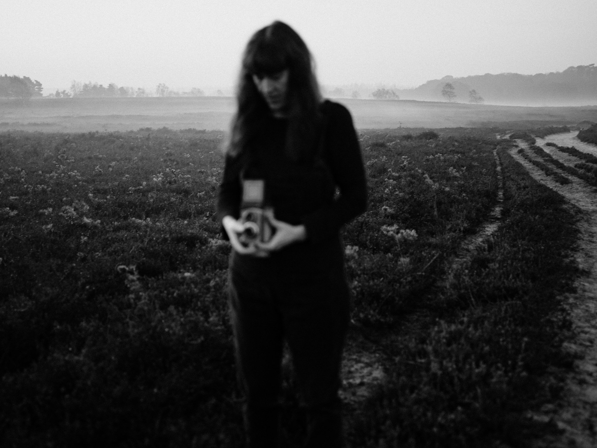 A woman with long dark hair, dressed in dark clothing, is standing in a field with tire tracks. She is looking down and holding an object. The background features rolling hills and a hazy sky, all in black and white.