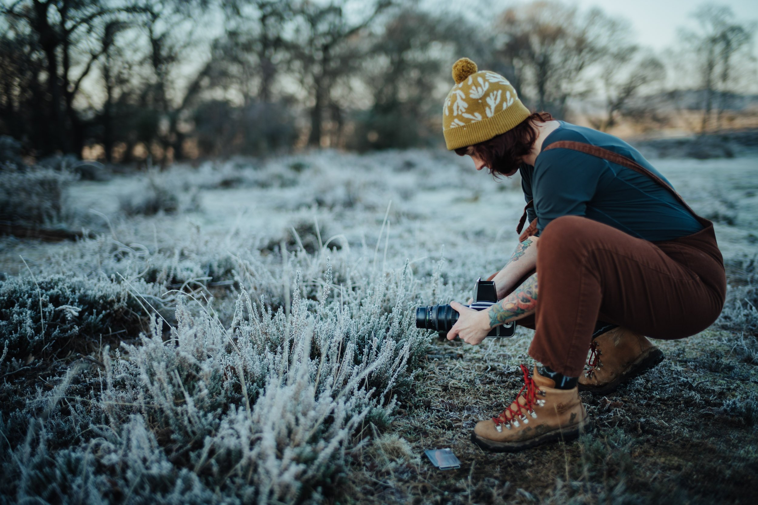 A person with tattoos wearing a yellow knit hat and brown boots, crouching down in a frost-covered field, holding a camera, with trees in the background.