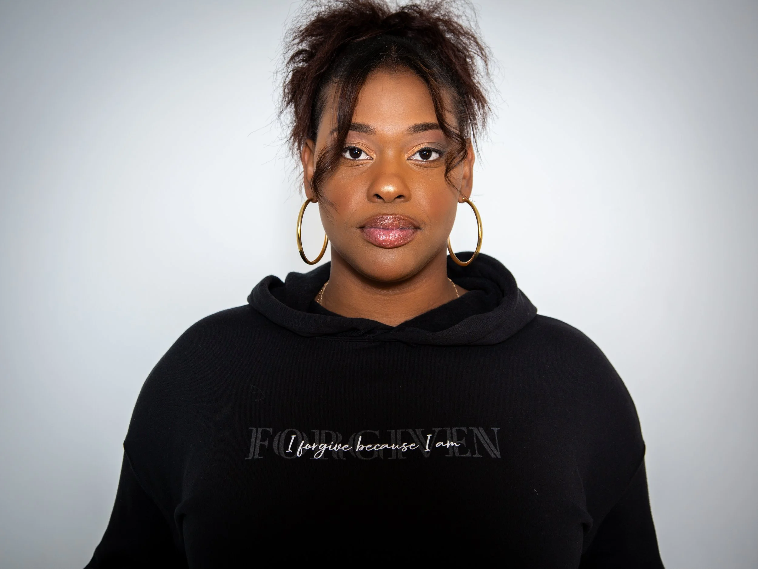 A woman with short curly hair, wearing large hoop earrings and a black hoodie, standing in front of a plain light gray background.