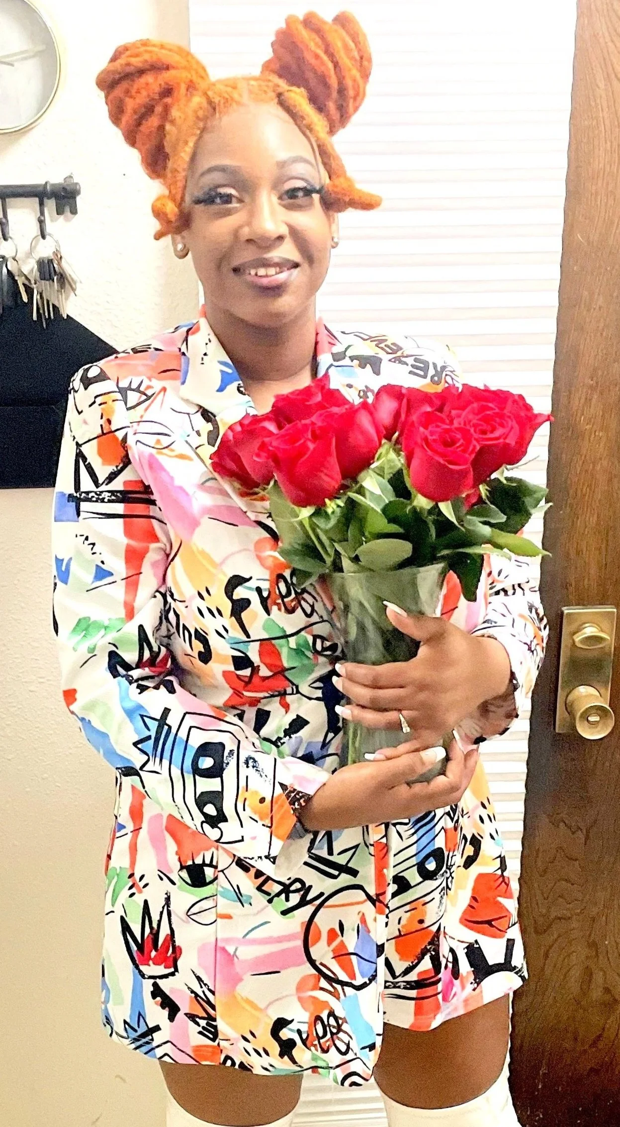 A woman with orange dreadlocks holding a bouquet of red roses, wearing a colorful graffiti-style patterned blazer, standing in front of a door.