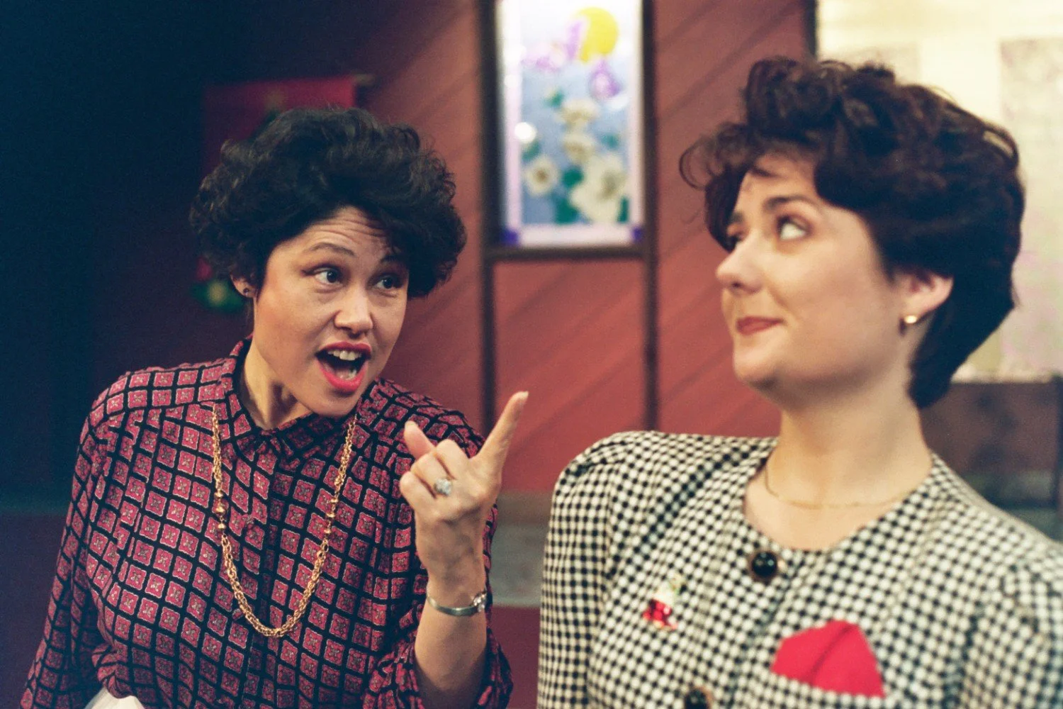 Two women indoors, one gesturing with a finger, wearing patterned tops, blurry background