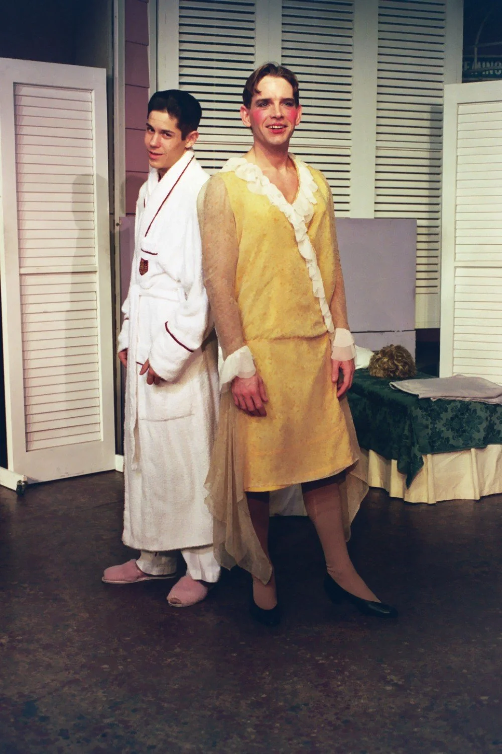 Two people posing indoors; one wearing a bathrobe and slippers, the other in a yellow dress with makeup.