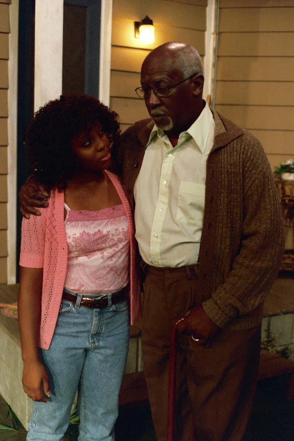 An elderly man and a young woman stand outside a house, sharing a warm moment. The man is wearing glasses, a brown cardigan, and is holding a cane, while the woman is wearing a pink top and blue jeans.