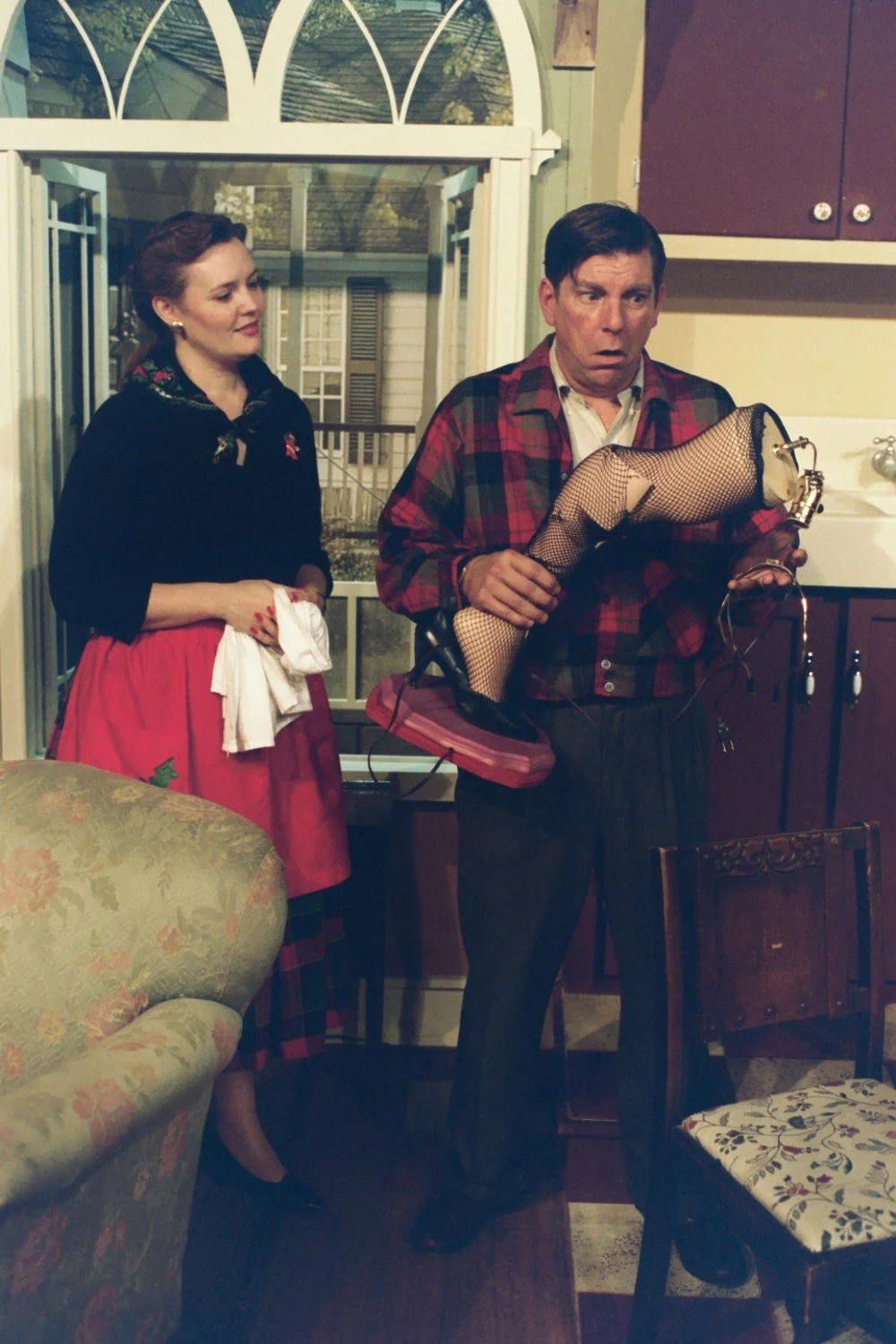 A man holding a novelty lamp shaped like a woman's leg wearing a fishnet stocking and high heel, accompanied by a woman in a vintage-style dress, set in a rustic interior.