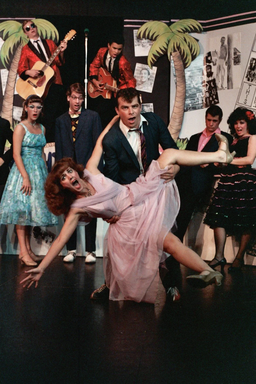 A group of people dressed in retro 1950s-style clothing performing on stage, with a man holding a woman in a dramatic dance pose. Background includes palm tree decorations and musicians with guitars.