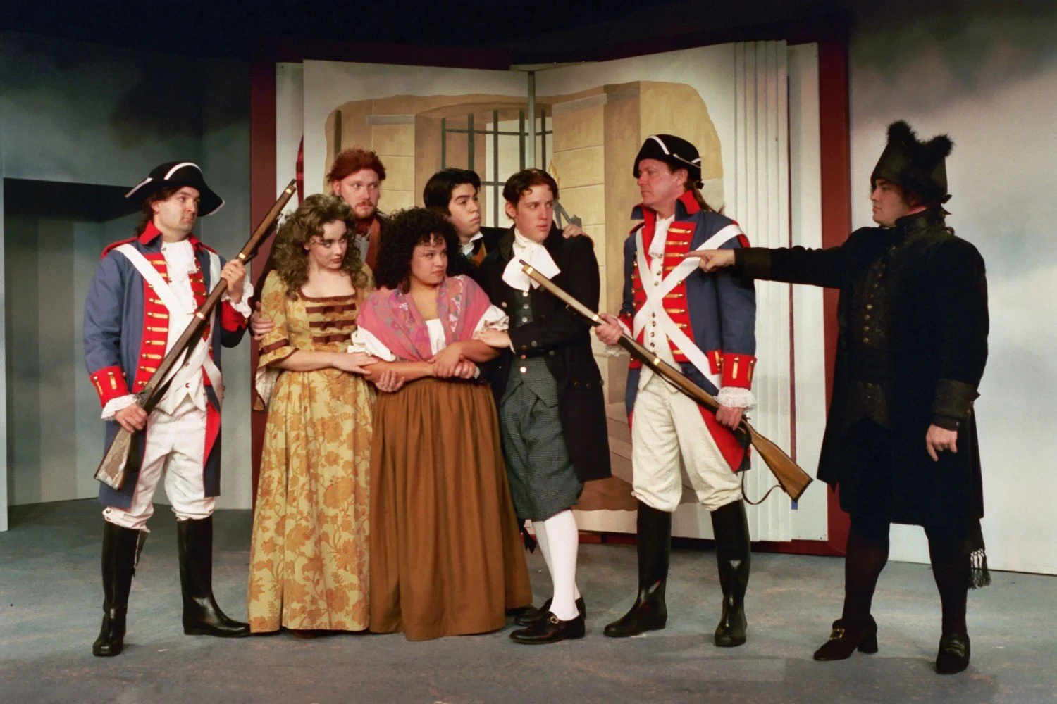 Group of actors in period costumes on stage, depicting a scene with soldiers holding muskets and individuals in historical attire. Backdrop features a painted setting.