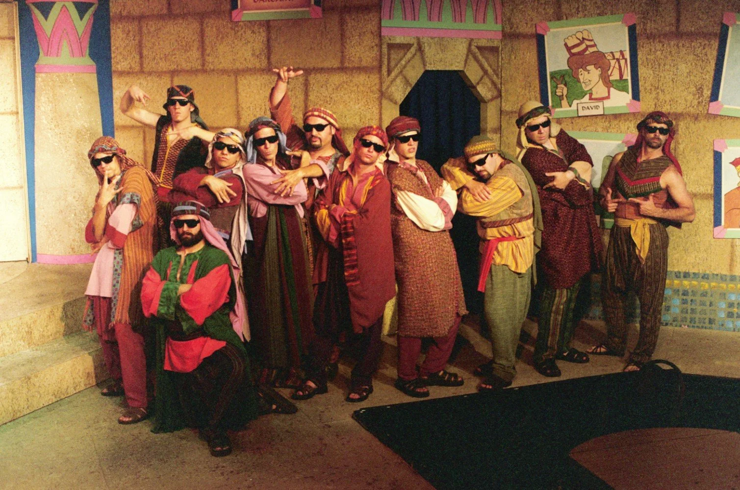 Group of people in colorful costumes and sunglasses posing on stage with Egyptian-themed backdrop.
