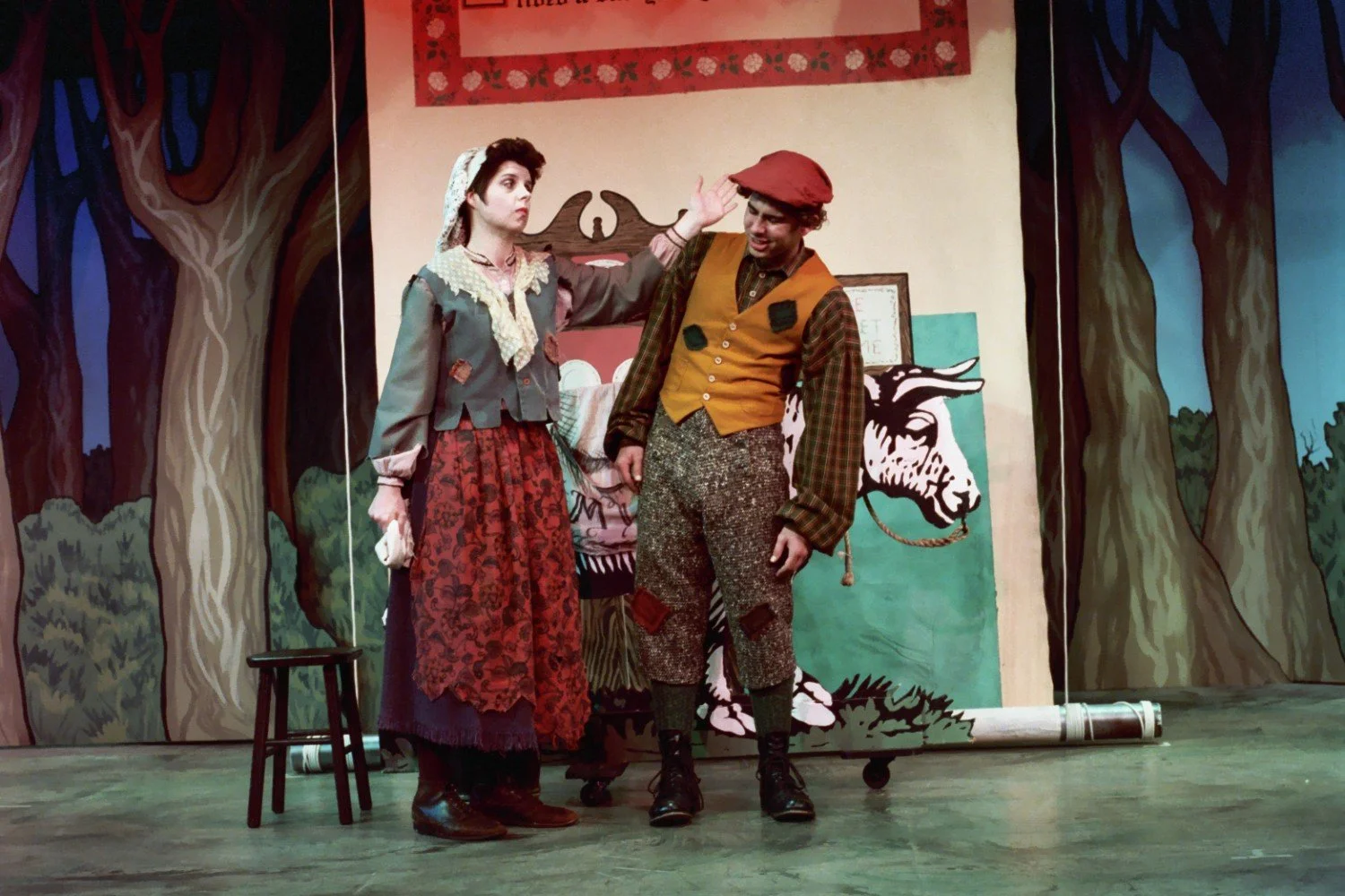 Actors in a theatrical performance wearing vintage costumes; one dressed as a peasant woman with a headscarf and long skirt, another as a man with a cap and patched vest, in front of a backdrop depicting trees and a cow illustration.