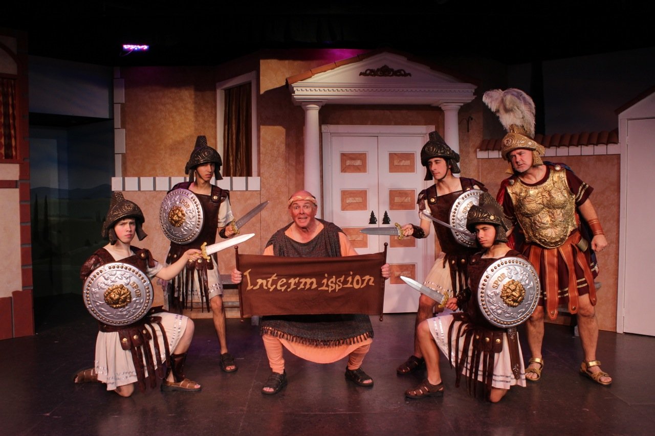 Stage performance with actors in Roman soldier costumes and a man holding an 'Intermission' sign.