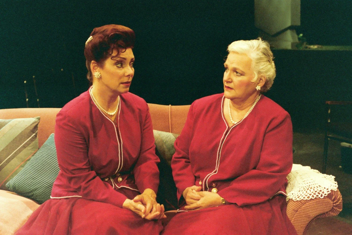 Two women in matching red outfits with pearls sitting on a couch, engaged in conversation, against a dark background.