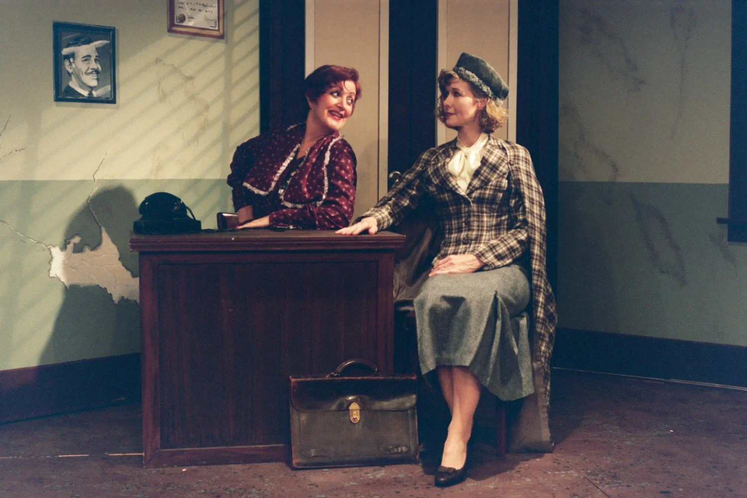 Two women dressed in vintage attire seated in an office-like setting, one behind a desk with a telephone and the other beside, against a wall with a sketch of a man's portrait.