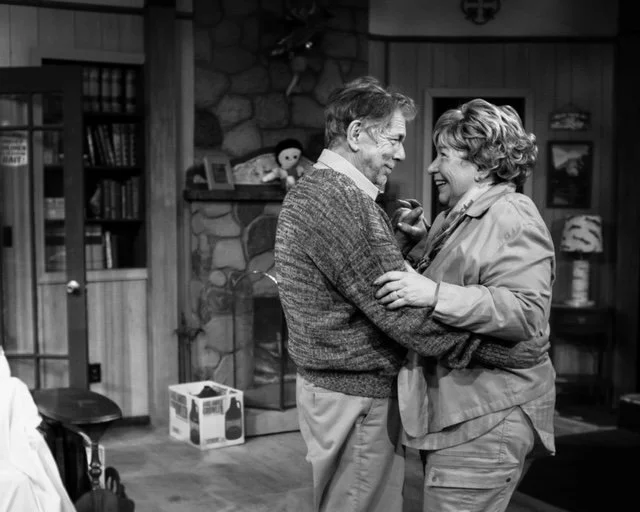 Older couple dancing in a cozy, rustic living room with stone fireplace and bookshelves, black and white photo.