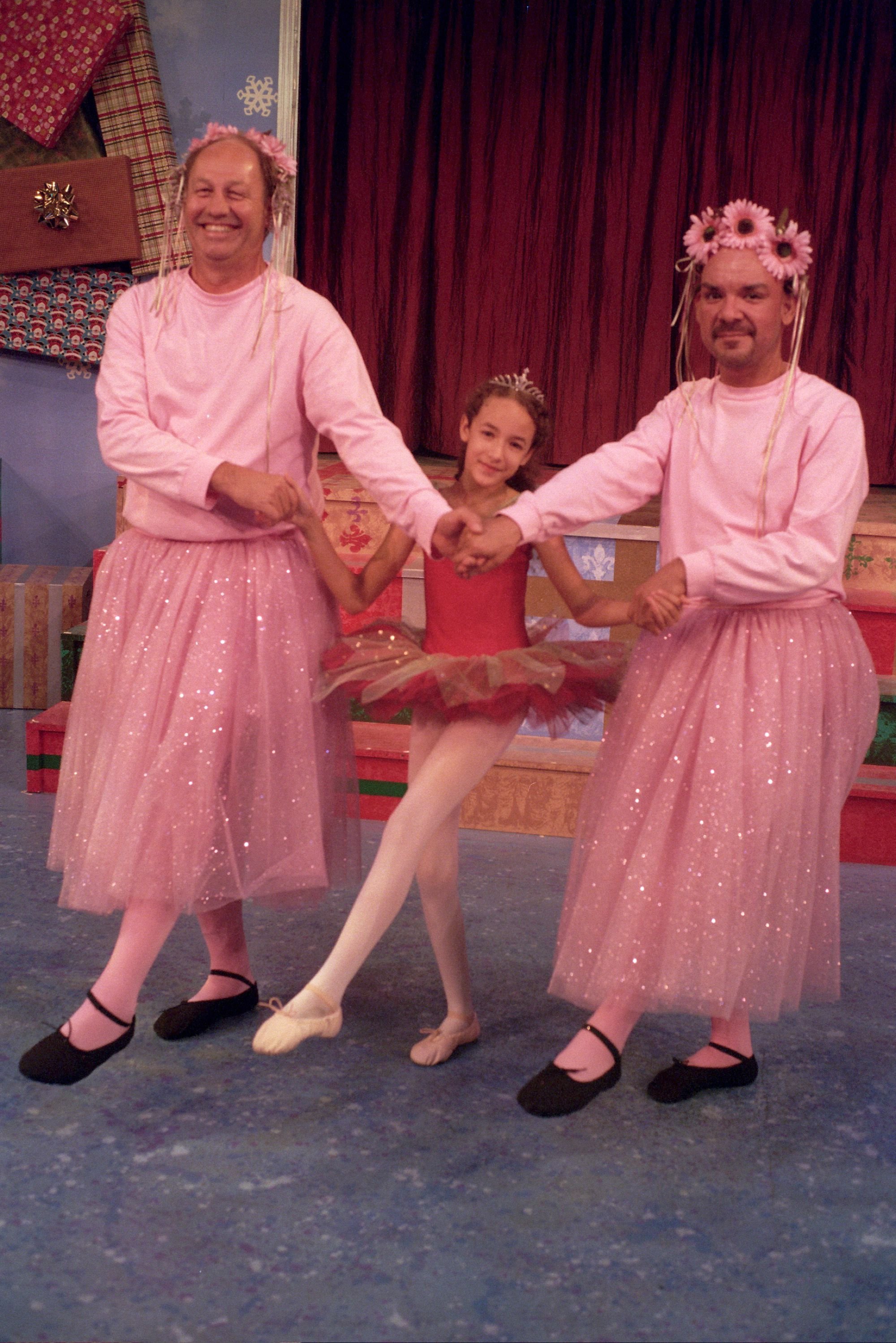 Two men in pink tutus and wigs pose with a young ballerina in a red costume on a stage.