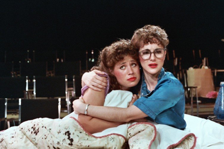Two women sitting closely together, embracing each other, both with concerned expressions. They are seated on a white surface with a patterned blanket, and the background is filled with empty chairs, suggesting a theater or studio setting.