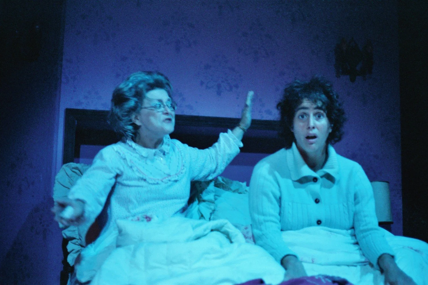 Two people in a dramatic scene on a stage, sitting up in bed with surprised and expressive gestures. The setting is dimly lit with a bluish hue.