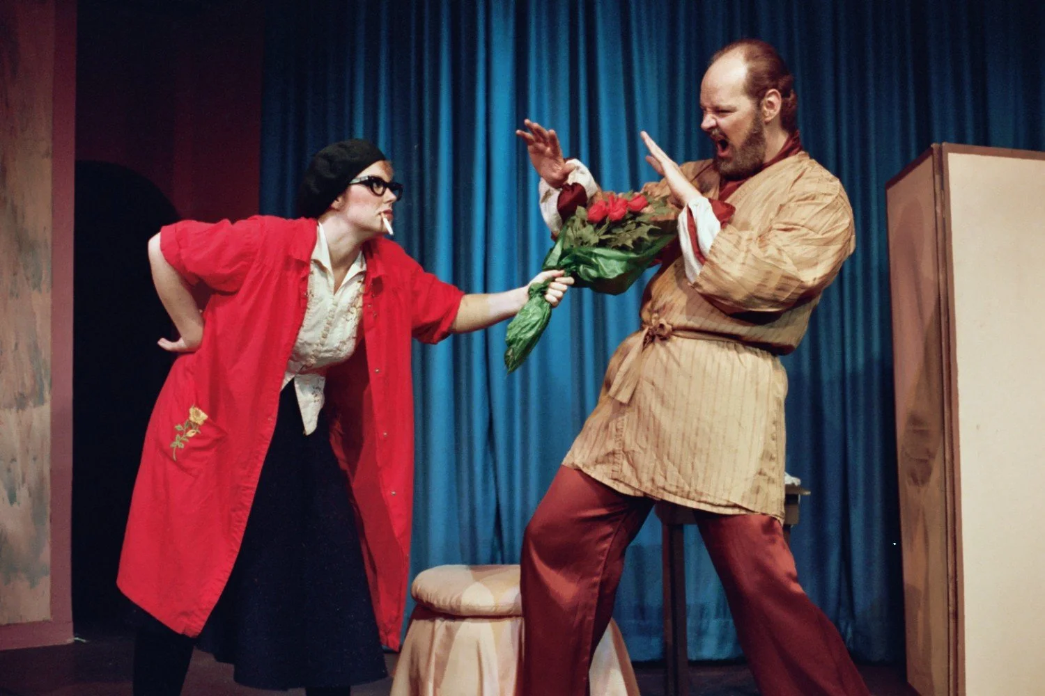 Two actors performing a comedic scene on stage. One is offering a bouquet of flowers, and the other is reacting with exaggerated surprise. They are wearing theatrical costumes with a blue curtain backdrop.