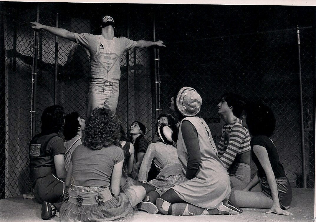 A group of nine people in theatrical costumes in front of a wire fence, with one person standing and the others sitting or kneeling, looking up at the standing person who has arms outstretched.